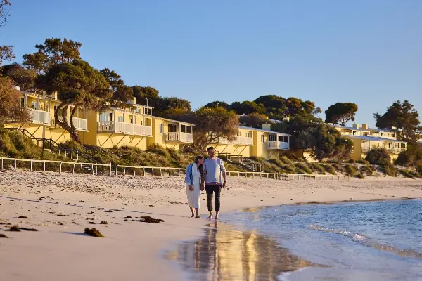 Couple holding hands walking along the beach with yellow Stay Rottnest self-contained accommodation in background