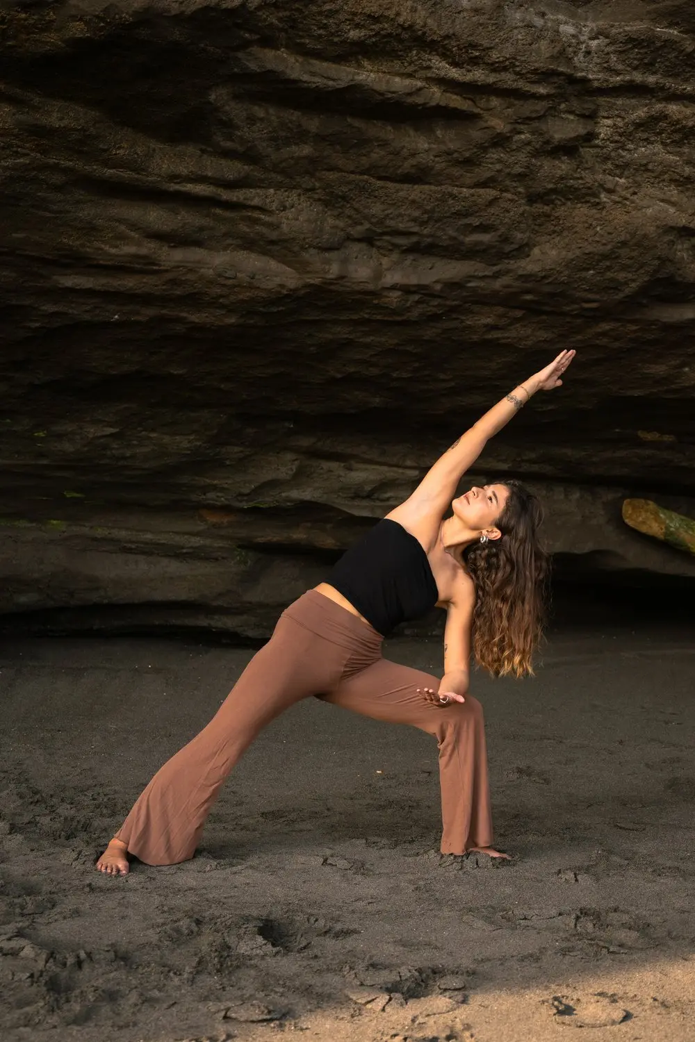 A woman practicing yoga poses on the sandy beach beneath a rocky overhang.