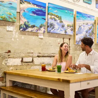 Couple eating at Geordie's Cafe & Art Gallery with art prints on wall behind them of Rottnest Island beaches